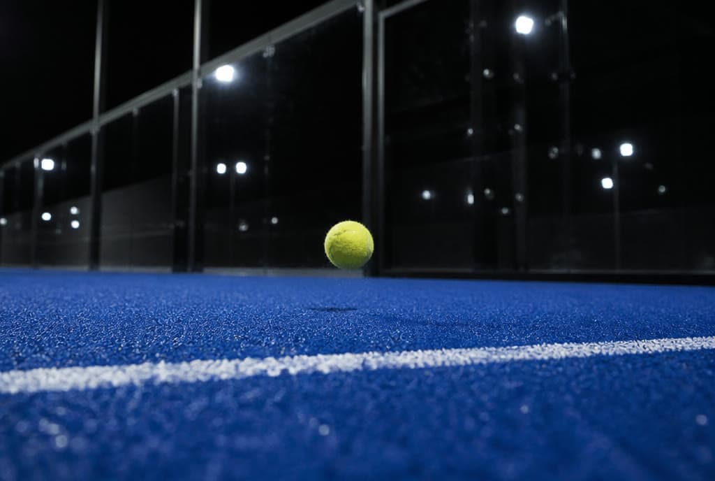 Una bola de pádel botando junto a la línea de saque de una pista azul, de noche