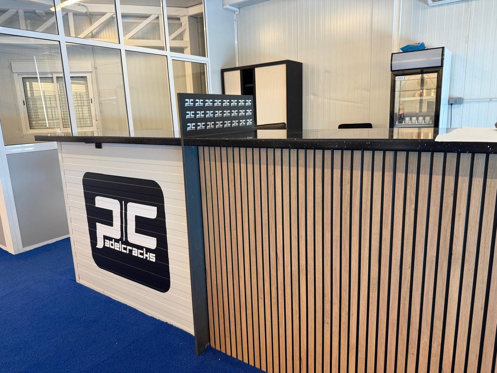 El mostrador de recepción, de listones de madera con encimera negra, con el logotipo de Pádel Cracks en la pared y la nevera de bebidas al fondo